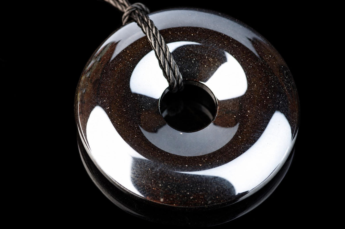 A polished, dark brown donut-shaped pendant with a central hole and speckled texture hangs from a twisted cord against a black background.