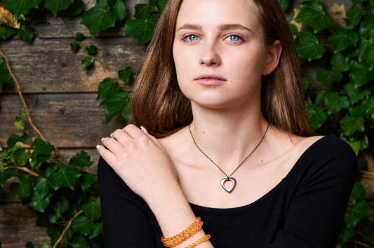 A woman in a black top wears a small heart-shaped hematite pendant on a dark cord, standing against a leafy wooden wall.