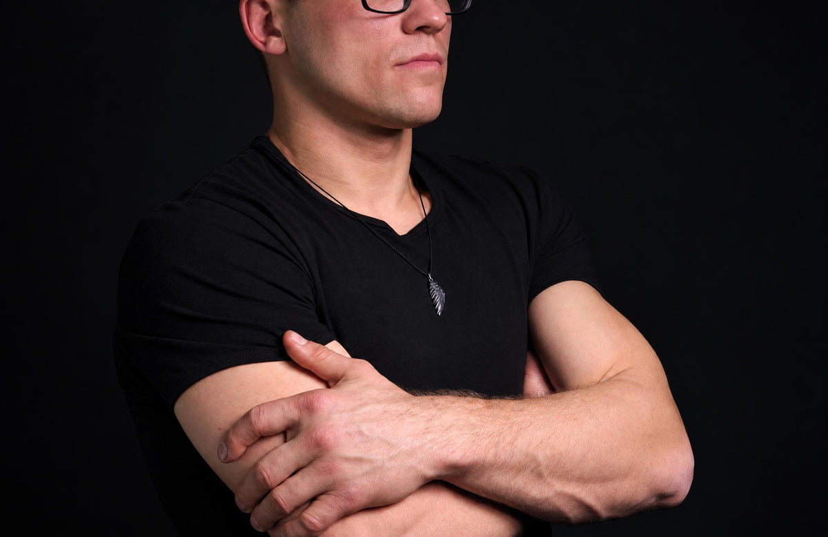 A man in a black t-shirt wears a silver-toned wing pendant on a dark cord against a dark surface.