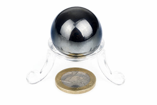 A polished reflective grey sphere sits on a clear stand above a coin on a white surface.