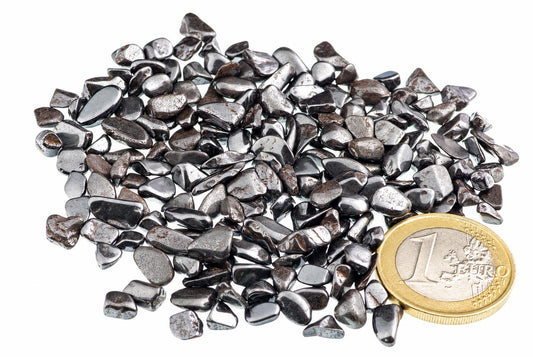 A pile of small, dark grey and silver-toned Hematite stones on a white surface with a one Euro coin for scale.