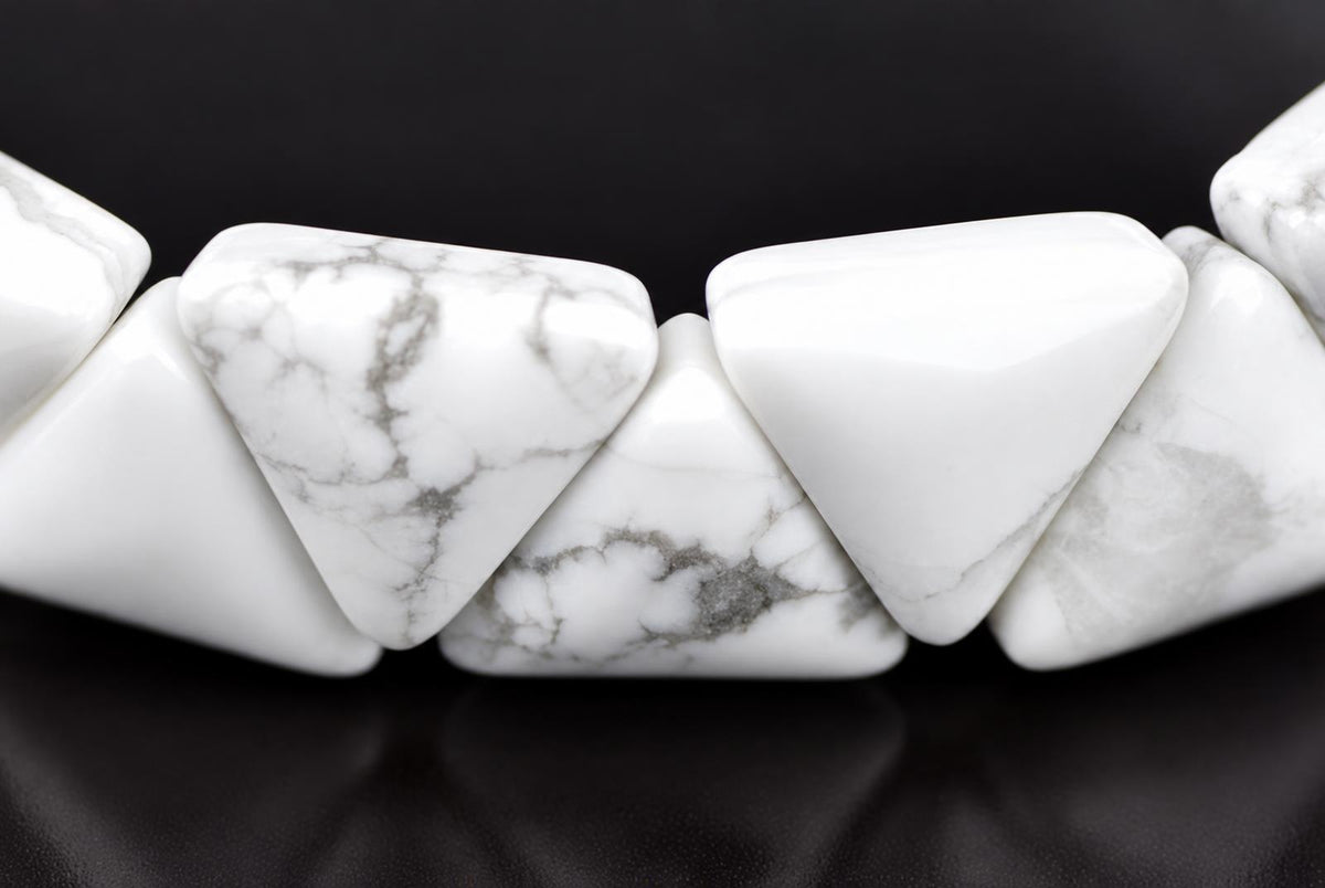 A close-up of a white and grey patterned Howlite bracelet with triangular beads on a dark surface.