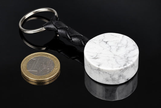 A white howlite keychain with grey veins, a black braided strap and a metal ring rests beside a coin on a dark surface.