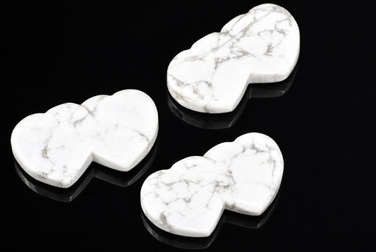 Three white Howlite pendants with grey veining, shaped like two joined hearts, on a dark reflective surface.