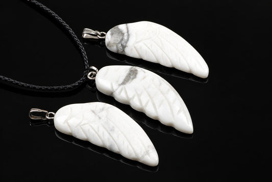 White howlite wing pendants with grey veins, carved feather grooves and silver-tone loops lie beside black cords on a glossy dark surface.