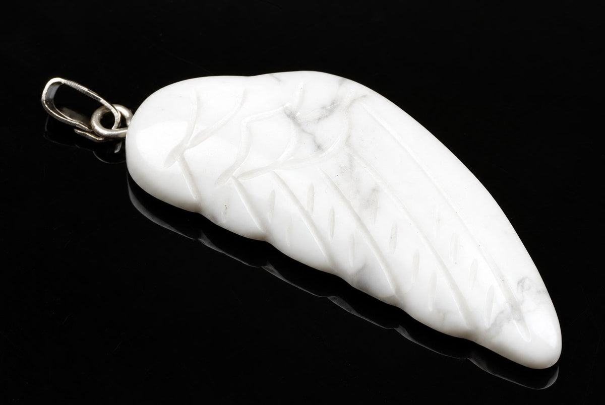 A white Howlite pendant shaped like a wing with carved veins, set in silver-toned metal on a dark surface.