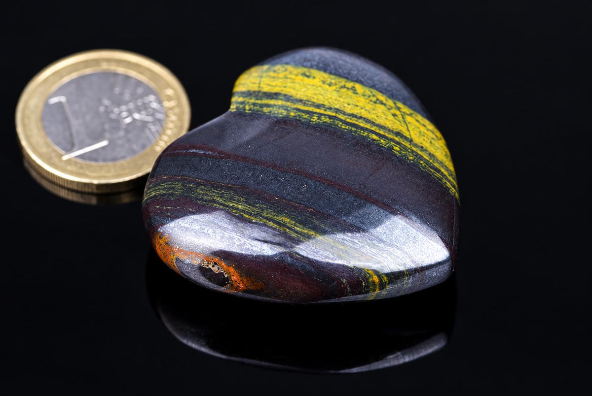 A polished, heart-shaped stone with dark bands and yellow stripes sits on a dark surface next to a coin.