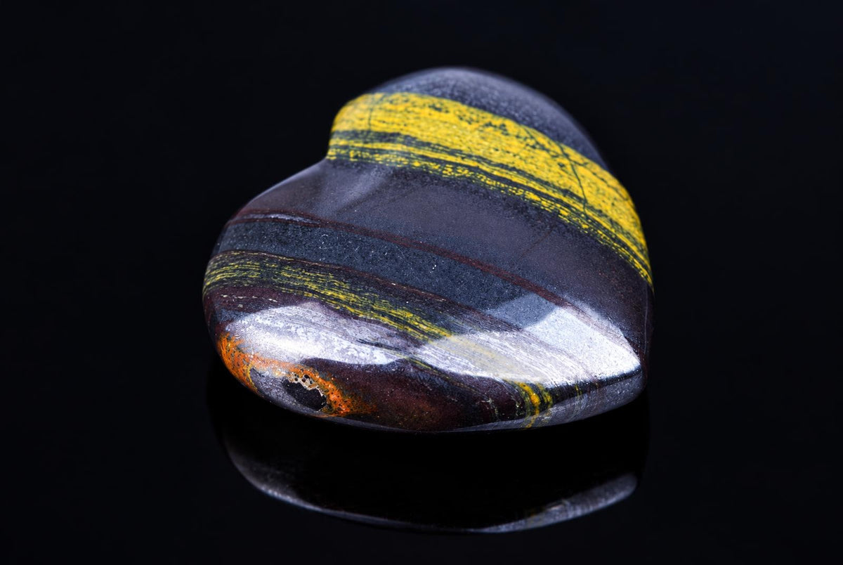A polished, heart-shaped stone with dark bands and bright yellow stripes on a black reflective surface.