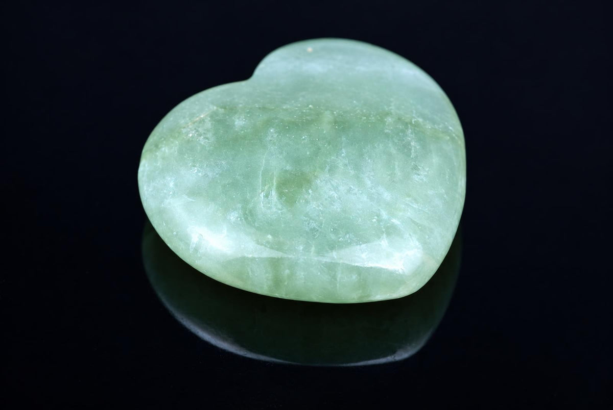 A polished, heart-shaped green stone rests on a dark, reflective surface.