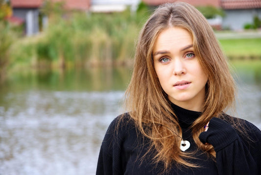 A woman with long brown hair wears a black turtleneck and a white heart-shaped pendant necklace against a blurred outdoor background.