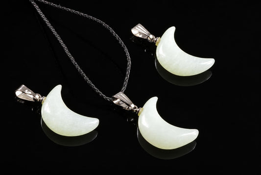 Pale green crescent moon jade pendants with silver-tone loops and black cords are arranged together on a glossy dark surface.