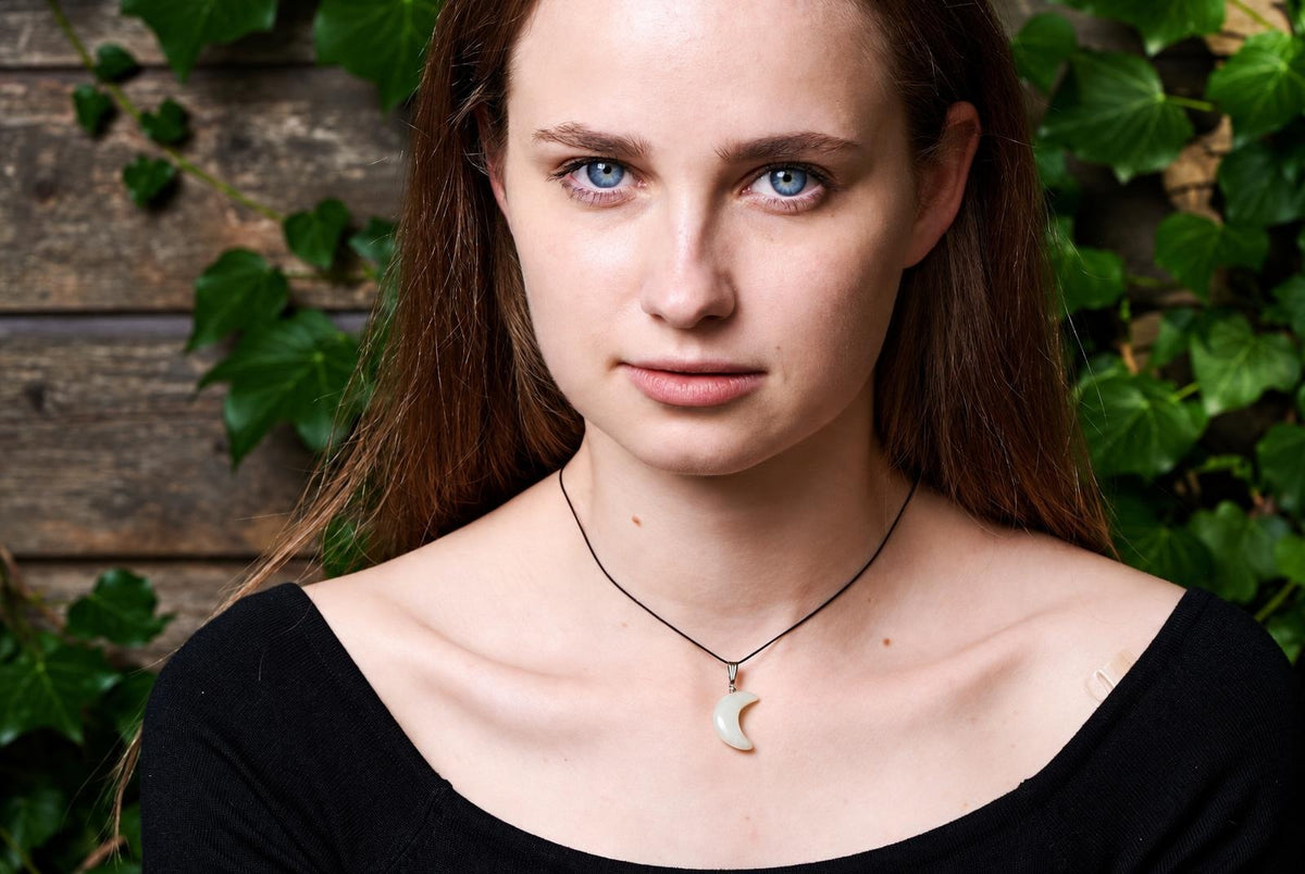 A woman wears a black top and a necklace with a pale, crescent-shaped pendant on a dark cord against a wooden background.