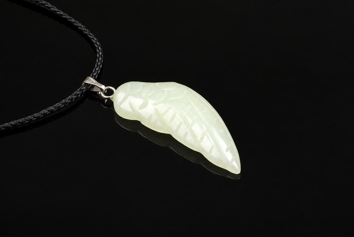 A pale green jade pendant carved into a wing shape with engraved lines, attached to a black cord via a silver-toned bail on a dark surface.