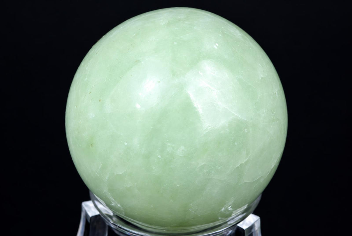 A polished, pale green spherical stone rests on a clear stand against a dark background.