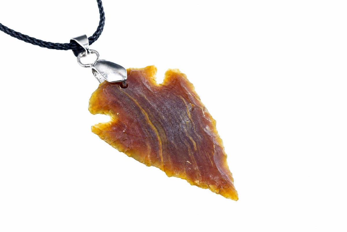 A brown and orange jasper arrowhead pendant shows layered bands and notched edges, suspended from a black cord on white.