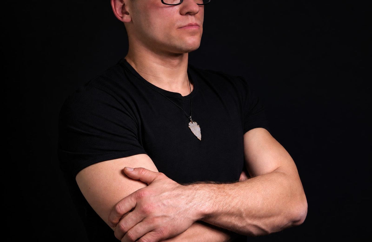 A man in a black t-shirt wears a pendant shaped like an arrowhead, light grey on a dark surface.