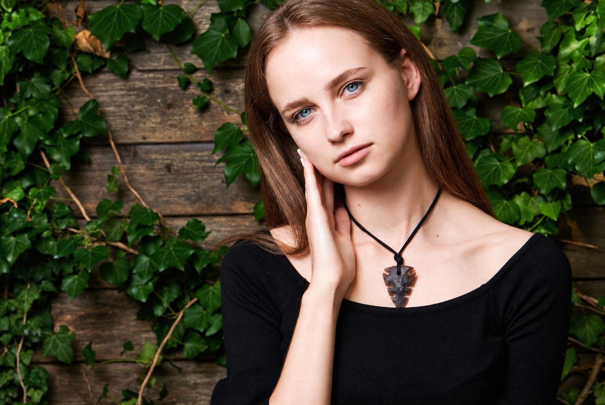 A woman wears a dark brown arrow-shaped pendant on a black cord against a wooden wall with green ivy.
