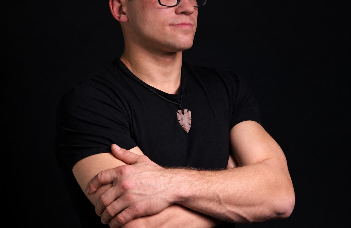 A man in a black t-shirt wears a light brown, arrow-shaped pendant on a dark cord against a dark background.