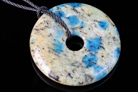 A polished, donut-shaped pendant with a central hole, featuring blue and black speckles on a pale surface, threaded with a grey cord against a dark background.