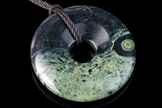 A polished, donut-shaped pendant with swirling green and dark patterns on a black surface, threaded with a dark cord.