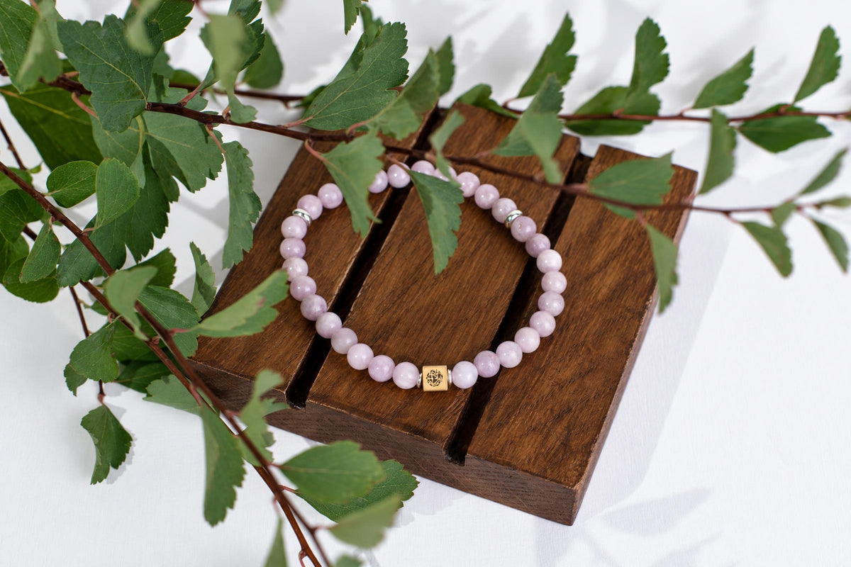 A bracelet with pale pink beads and a gold-toned square charm rests on a dark wooden surface, surrounded by green leaves.