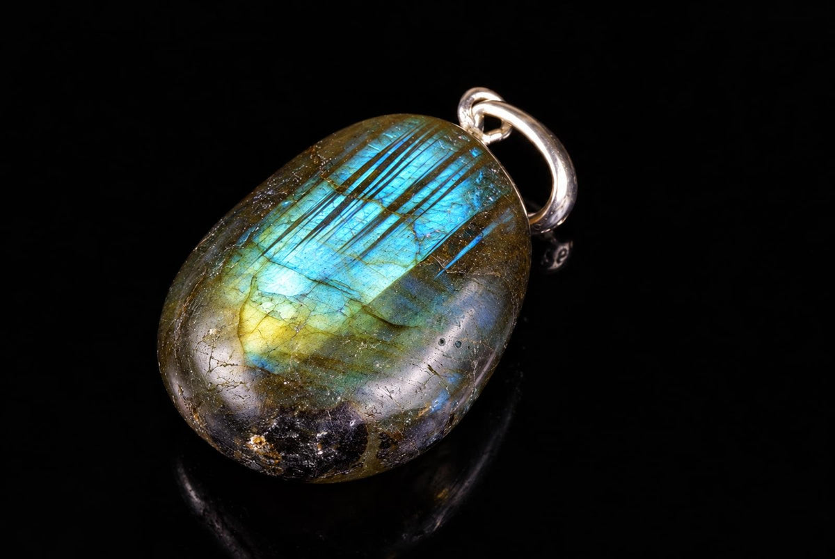 A polished oval labradorite pendant with blue and yellow flashes, set in a silver-toned loop bail on a dark reflective surface.