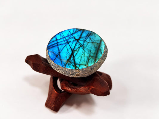A polished oval labradorite stone with blue and green iridescence sits on a dark wooden stand against a white background.