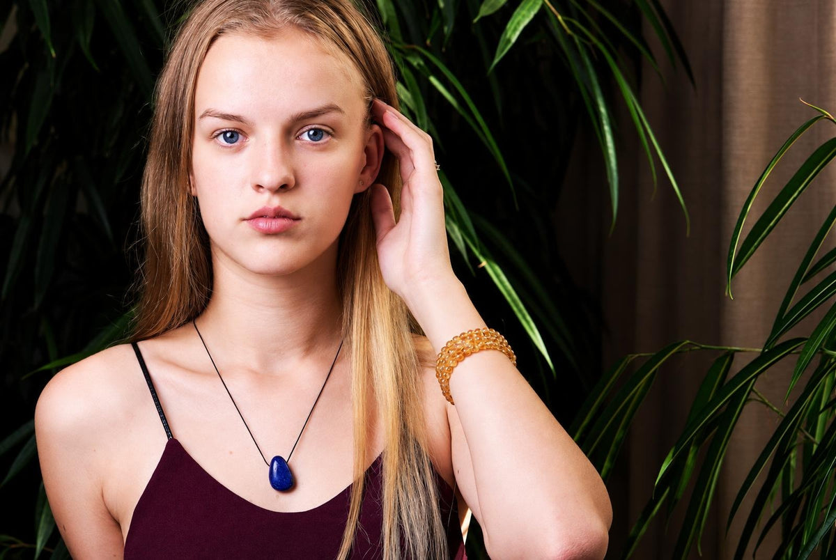A woman wears a dark blue teardrop pendant on a black cord and a gold-coloured beaded bracelet, with green plants in the background.