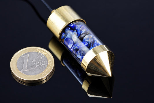A brass pendulum with a clear tube filled with blue and white stones, resting on a dark reflective surface next to a one euro coin.