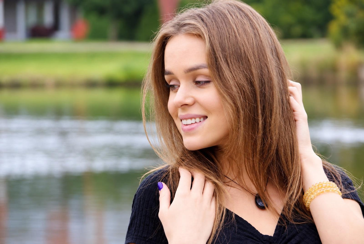 A smiling woman with long brown hair wears a black top, a dark pendant necklace and a yellow beaded bracelet outdoors near water.