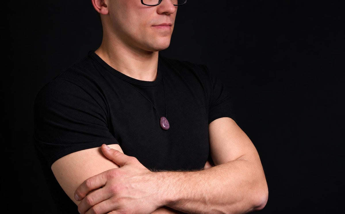 A man wears a black t-shirt with a dark purple, oval-shaped pendant on a thin cord against a dark background.