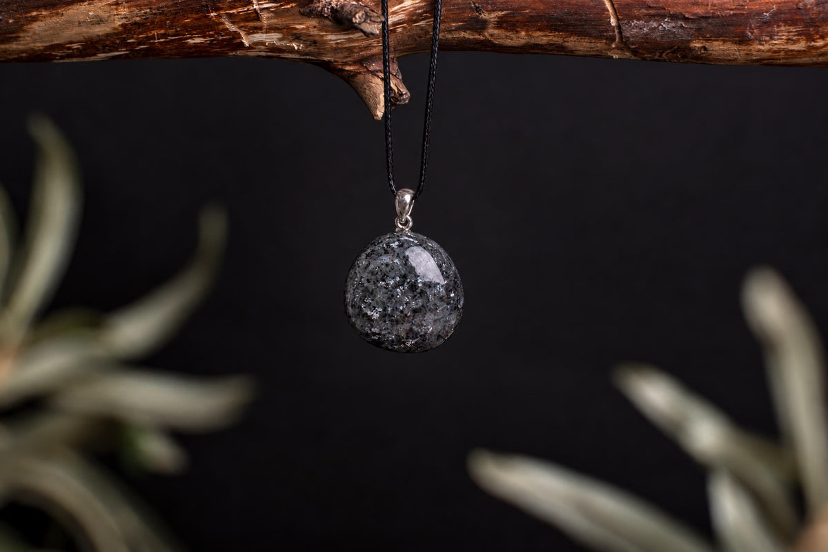 A round, polished Lithomanite pendant with a speckled grey and black surface hangs from a black cord on a silver bail.