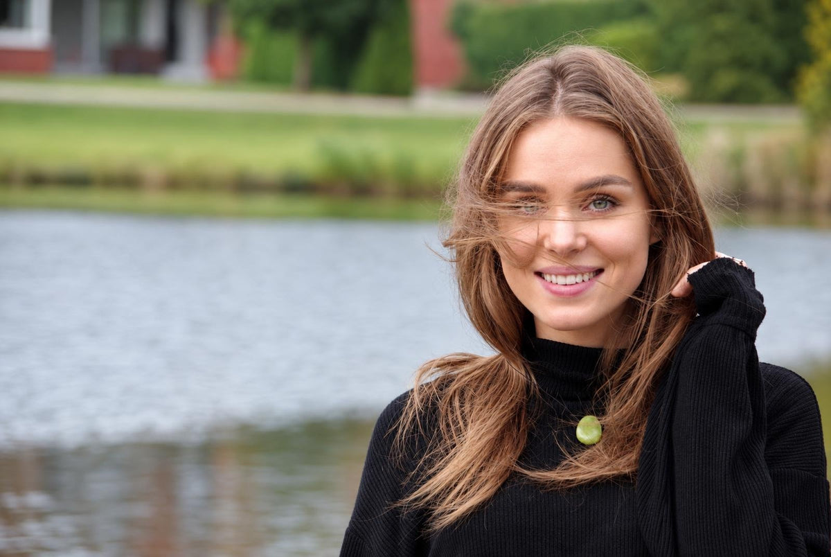A smiling woman with long brown hair wears a black turtleneck sweater featuring a round, light green pendant at the neckline.