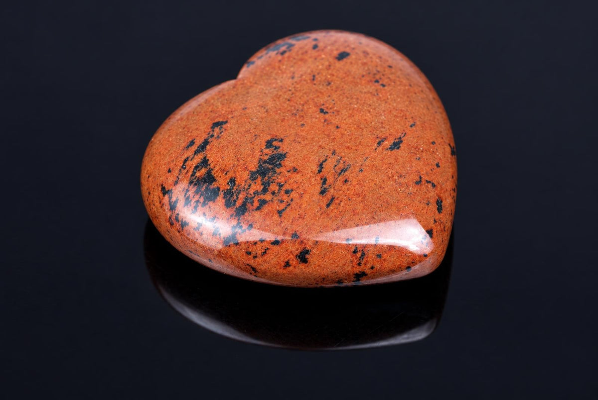 A polished, heart-shaped stone with a reddish-brown surface and black speckles rests on a dark, reflective surface.