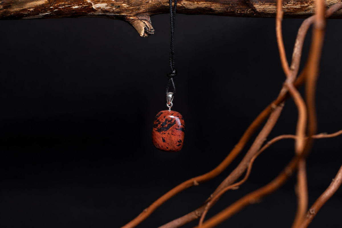 A polished red pendant with black speckles hangs from a black cord on a silver bail, suspended against a dark background near brown branches.