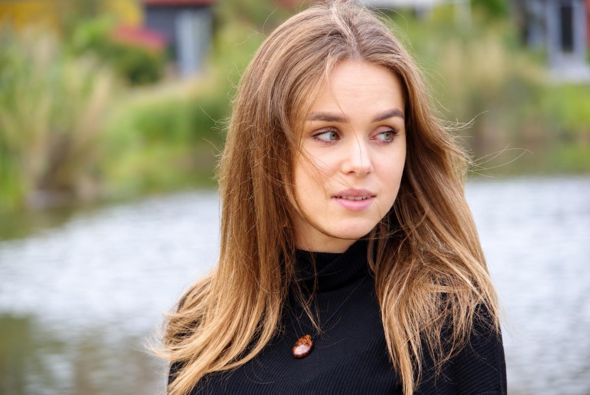 A woman with long brown hair wears a black turtleneck and a small, oval-shaped pendant with dark brown and lighter tones.