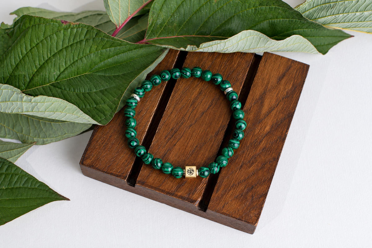 A green beaded bracelet with a gold-toned square charm rests on a dark wooden surface, surrounded by large green leaves.