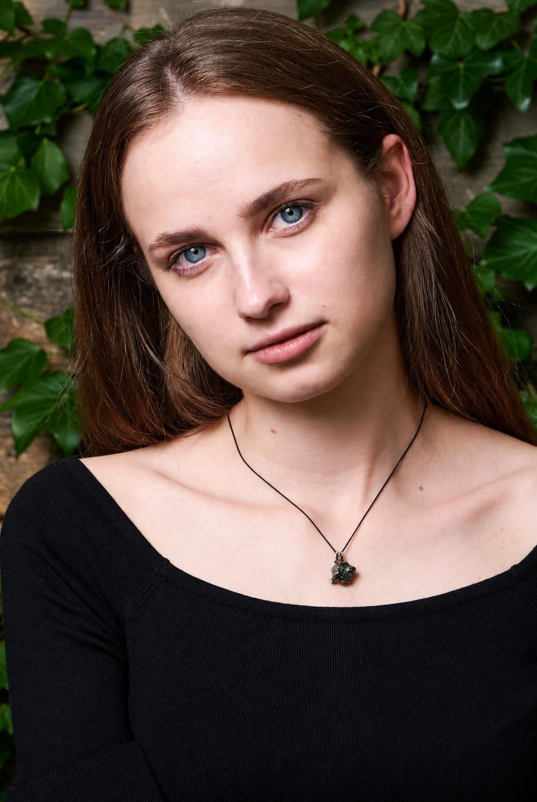 A woman wears a black top and a necklace with a dark, irregular pendant on a thin cord against a leafy background.