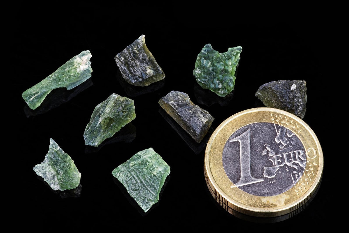 Several green and dark, translucent Moldavite fragments with rough shapes on a black surface next to a one Euro coin.
