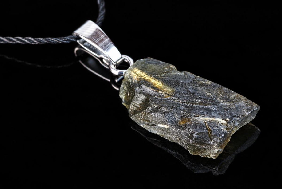 A dark green, rough-textured pendant with golden inclusions hangs from a silver-toned bail on a black cord against a dark surface.