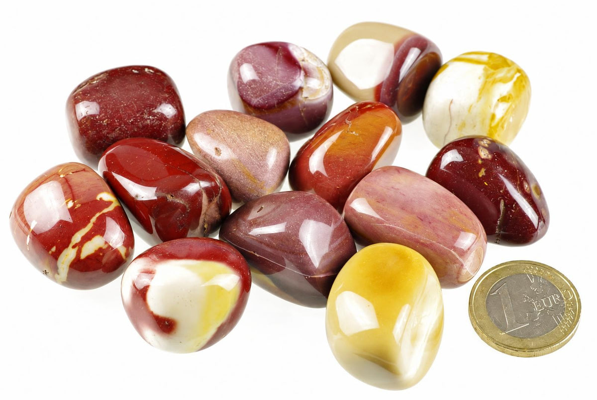 A collection of polished Mookaite Jasper stones in red, brown, yellow and white patterns on a pale surface with a one euro coin for scale.