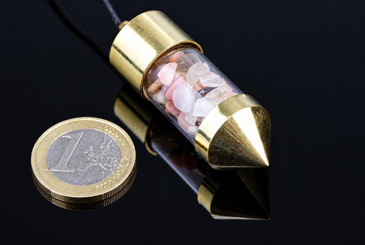 A brass pendulum with a clear tube containing pink and white stones, resting on a dark surface next to a one-euro coin.
