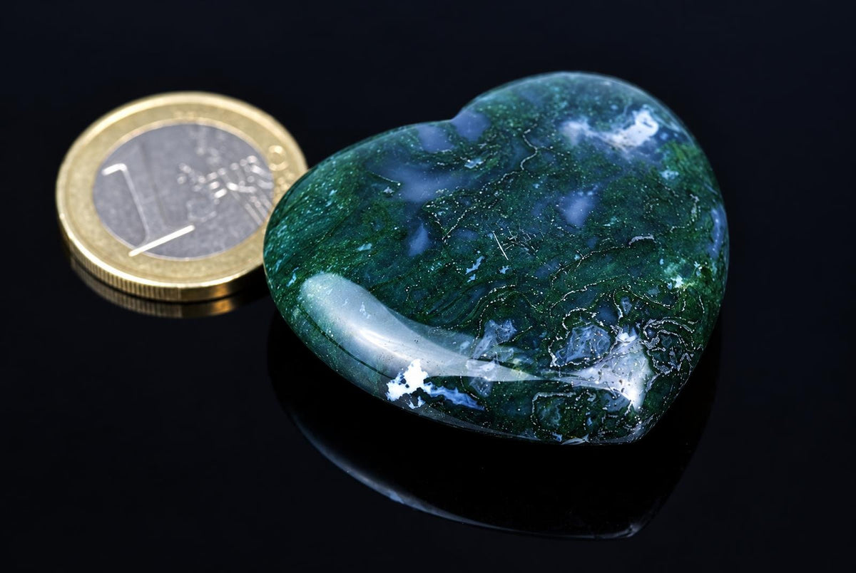 A polished dark green heart-shaped stone with pale cloudy patches rests beside a coin on a reflective black surface.