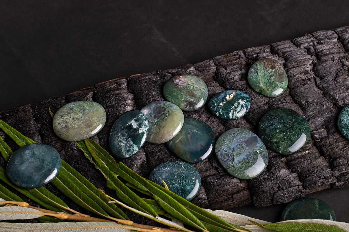 Polished green and blue moss agate palmstones arranged on a dark wooden surface with green leaves.