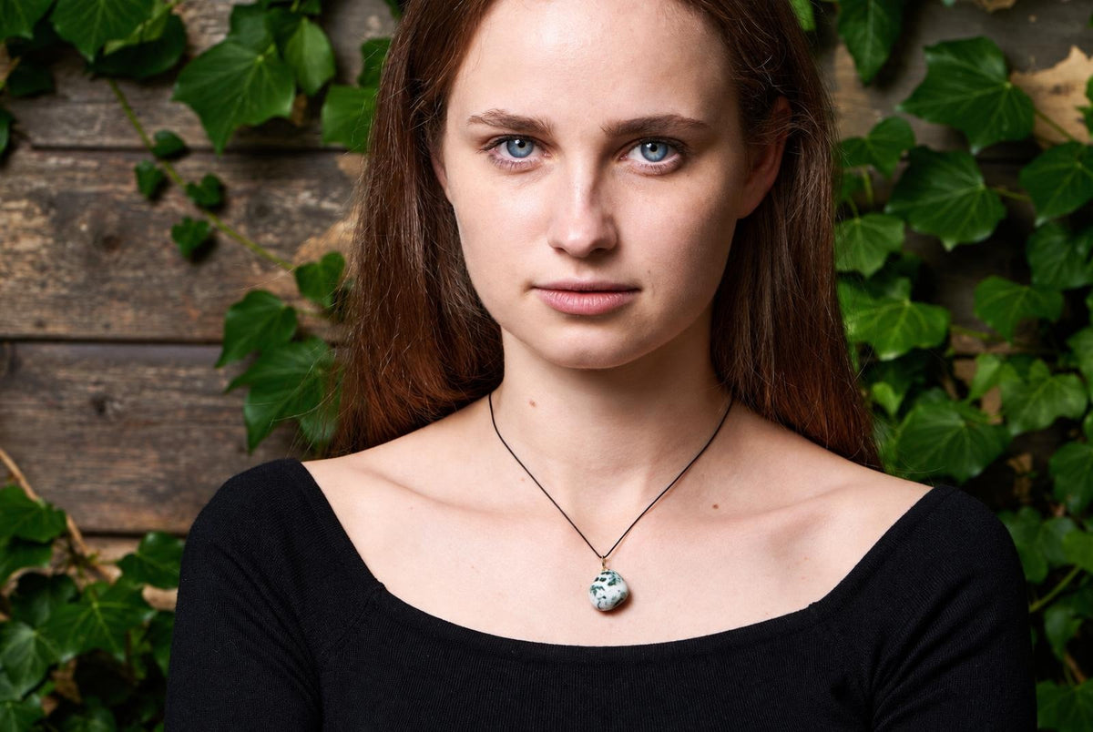 A woman wears a pendant with a white and green spotted stone on a thin black cord, set against a wooden background with ivy.