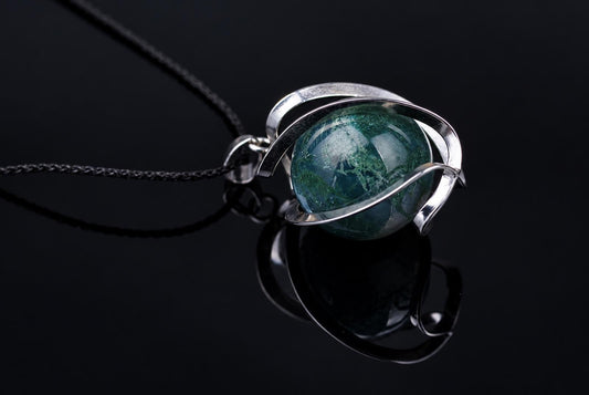A silver-toned pendant with a round, green and white patterned stone on a black cord, resting on a dark reflective surface.