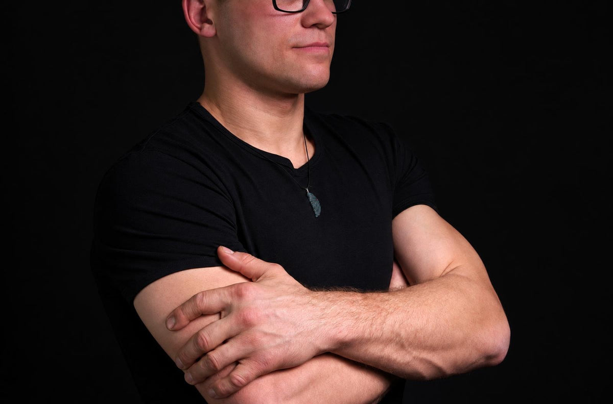 A man in a black t-shirt wears a dark green pendant on a thin cord against a dark background.