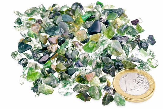 A pile of small, rough moss agate stones in various shades of green and white on a pale surface next to a one euro coin.