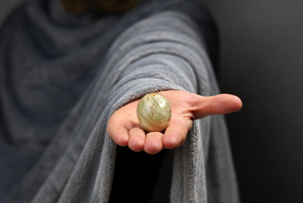 A hand holds a polished, oval-shaped stone with green and brown swirls against a dark background.