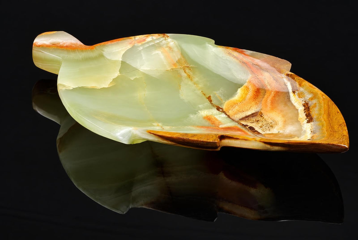 A polished onyx plate shaped like a leaf, featuring pale green and orange-brown bands, resting on a dark reflective surface.
